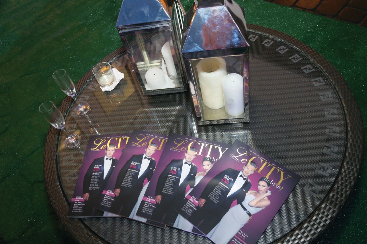 Copies of Le CITY deluxe fanned out on a glass table beside silver lanterns and champagne flutes at an evening event.