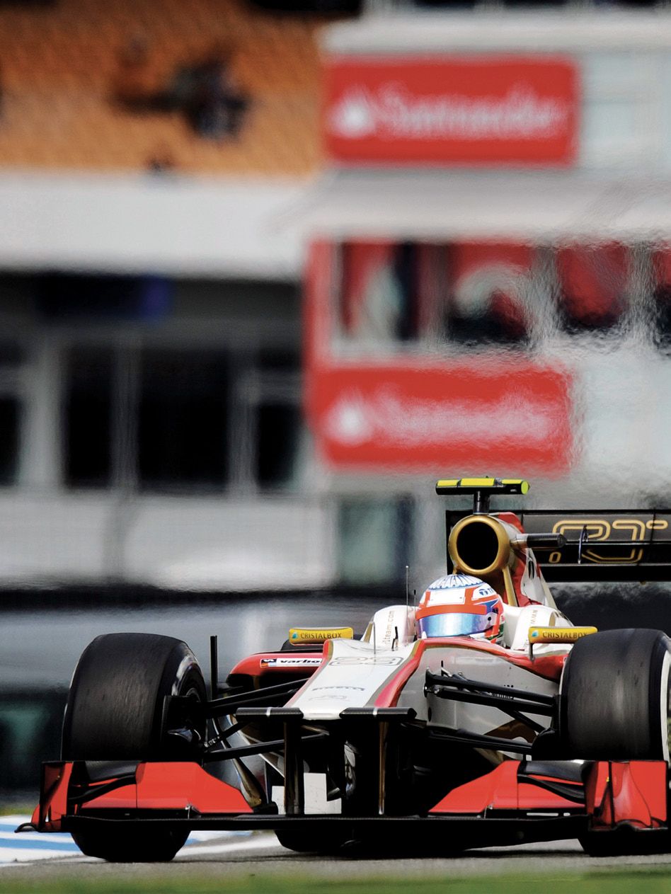 The red and white HRT Formula 1 car cornering on track, Santander banners blurred behind it.